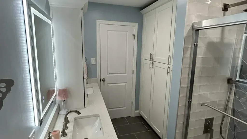 Bathroom with a tall white linen cabinet, a vanity with a lighted mirror, and a sliding glass shower