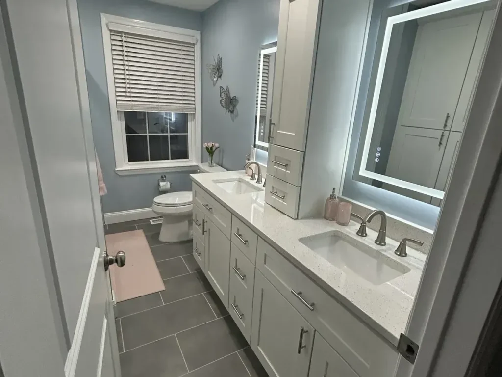 Long remodeled bathroom with a toilet by the window, a white vanity, a tall linen tower, and a lighted mirror