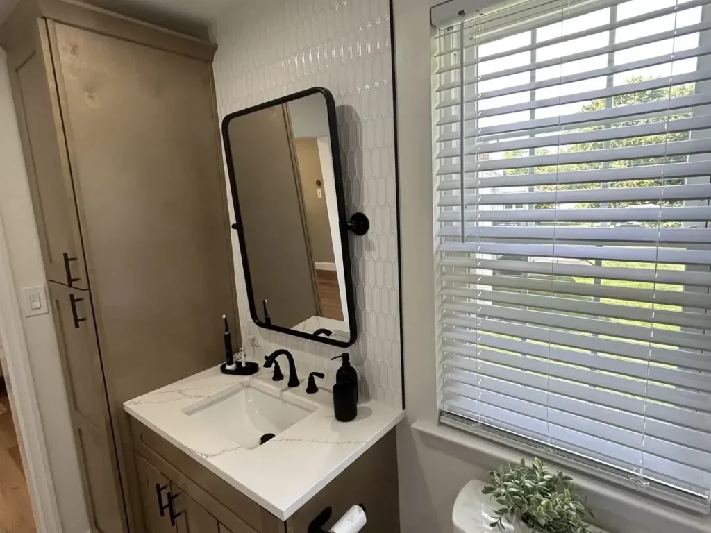 Small remodeled bathroom with a black framed pivot mirror, white vanity top, and tall wood cabinet by a window