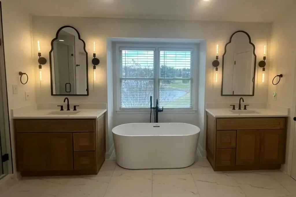 White freestanding soaking tub under a window between two wood vanities with arched black mirrors and sconces